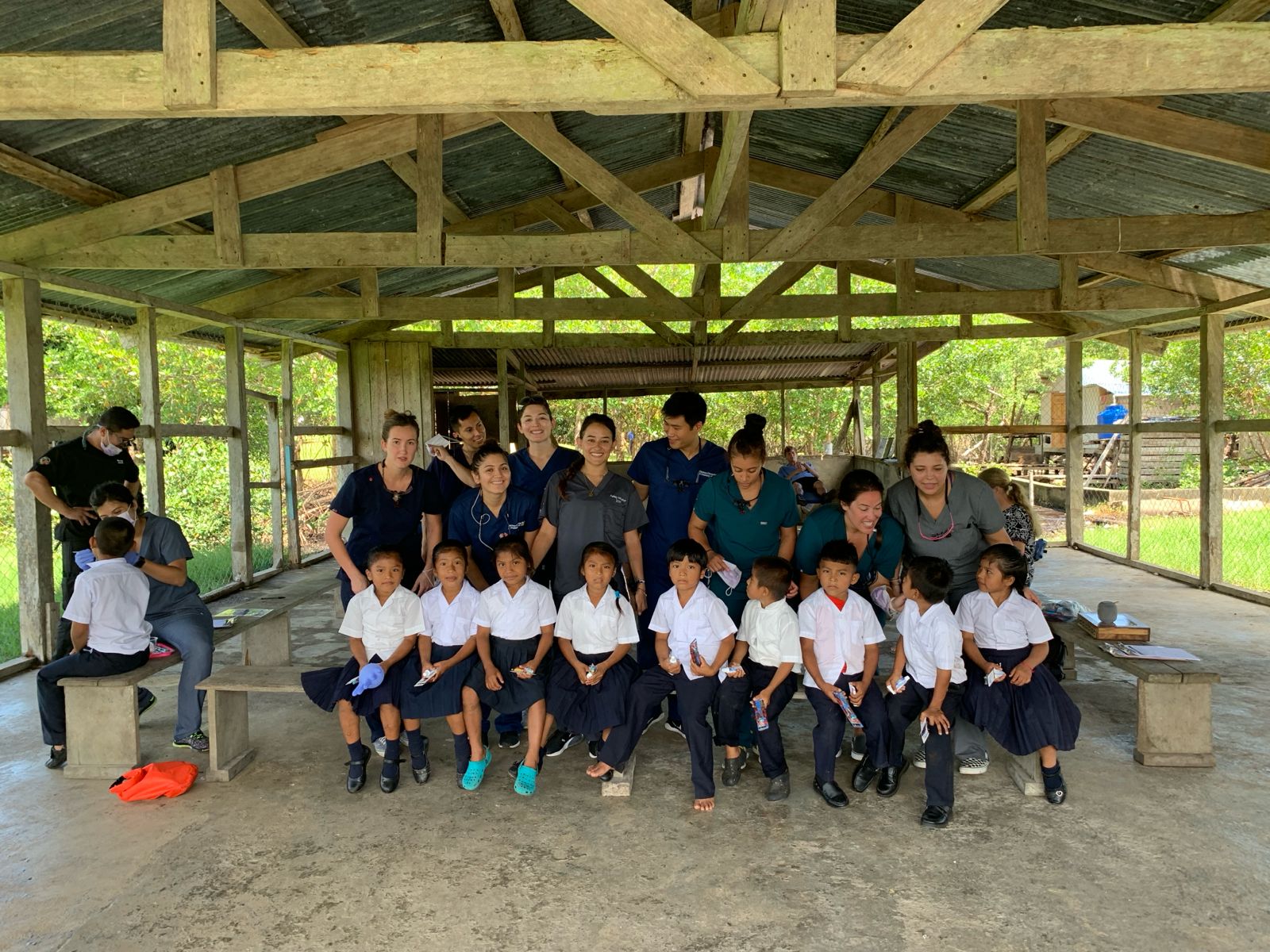 Schoolchildren in white shirts sit with volunteers under an open wooden shelter