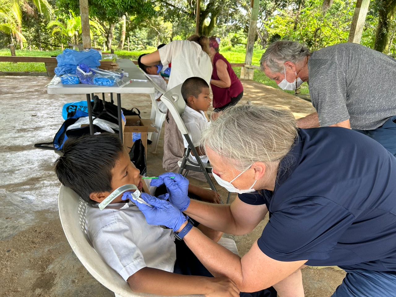 A volunteer in gloves and a mask treats a schoolboy outdoors while other children wait